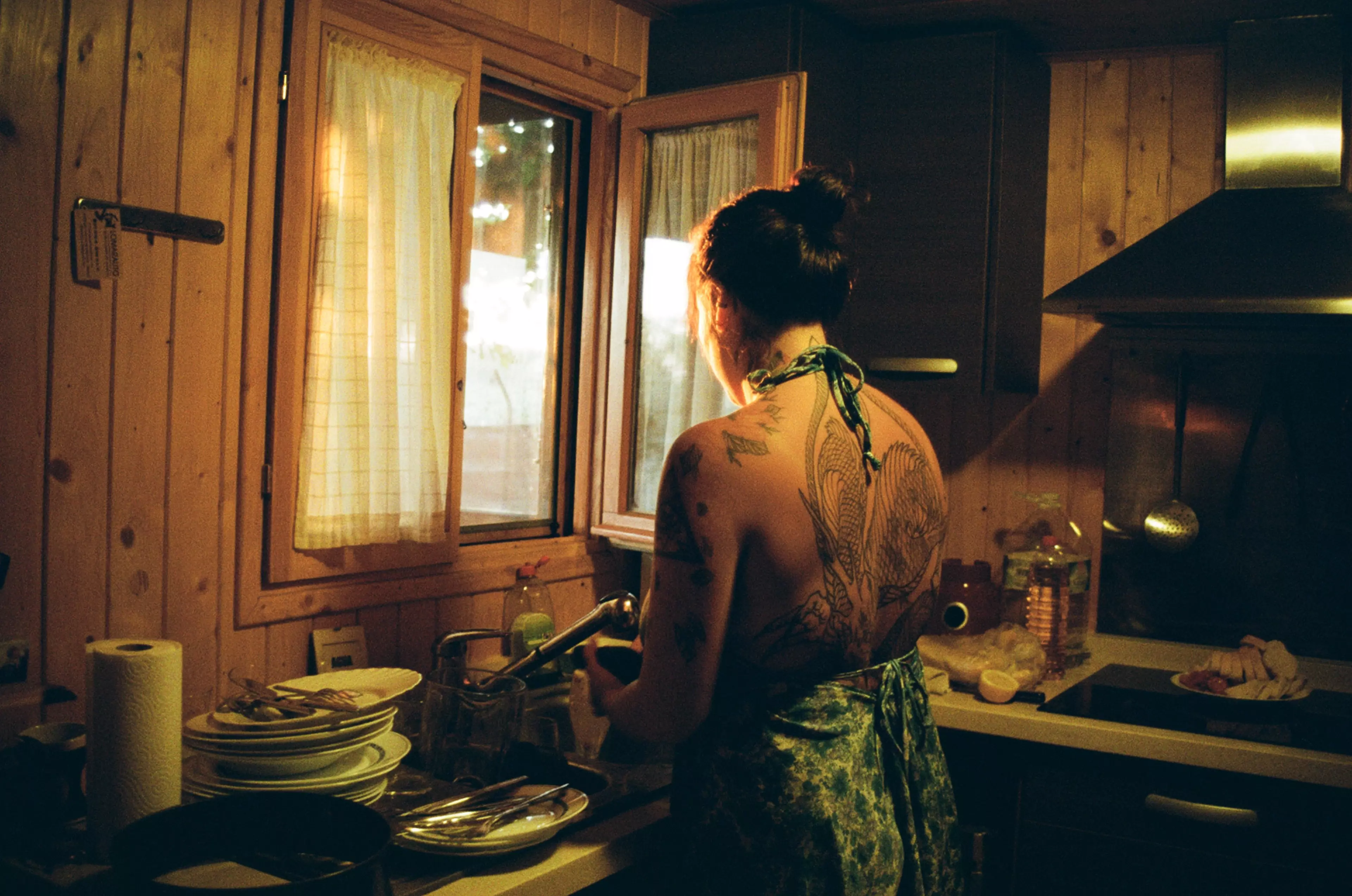 Dramatic photograph by Agostina Cerullo. A woman in a flowing summer dress stands in a kitchen, facing an open window. Her elaborate back tattoo is visible as she gazes outside.