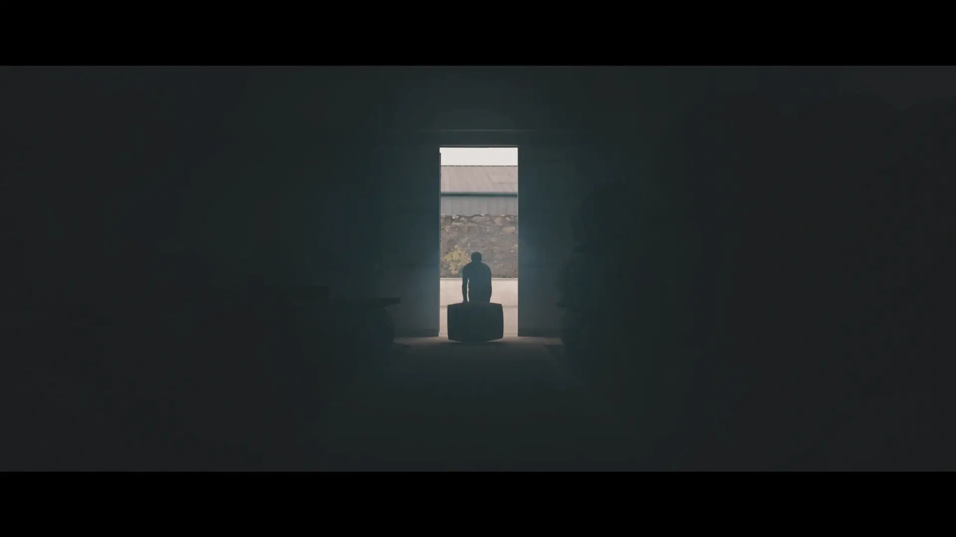 A scene from the 'KILCHOMAN: 100% ISLAY FROM BARLEY TO BOTTLE' film, with cinematography by Paolo Bischi. The image shows the silhouette of a man illuminated by light through an open door as he rolls a whiskey barrel into the distillery.