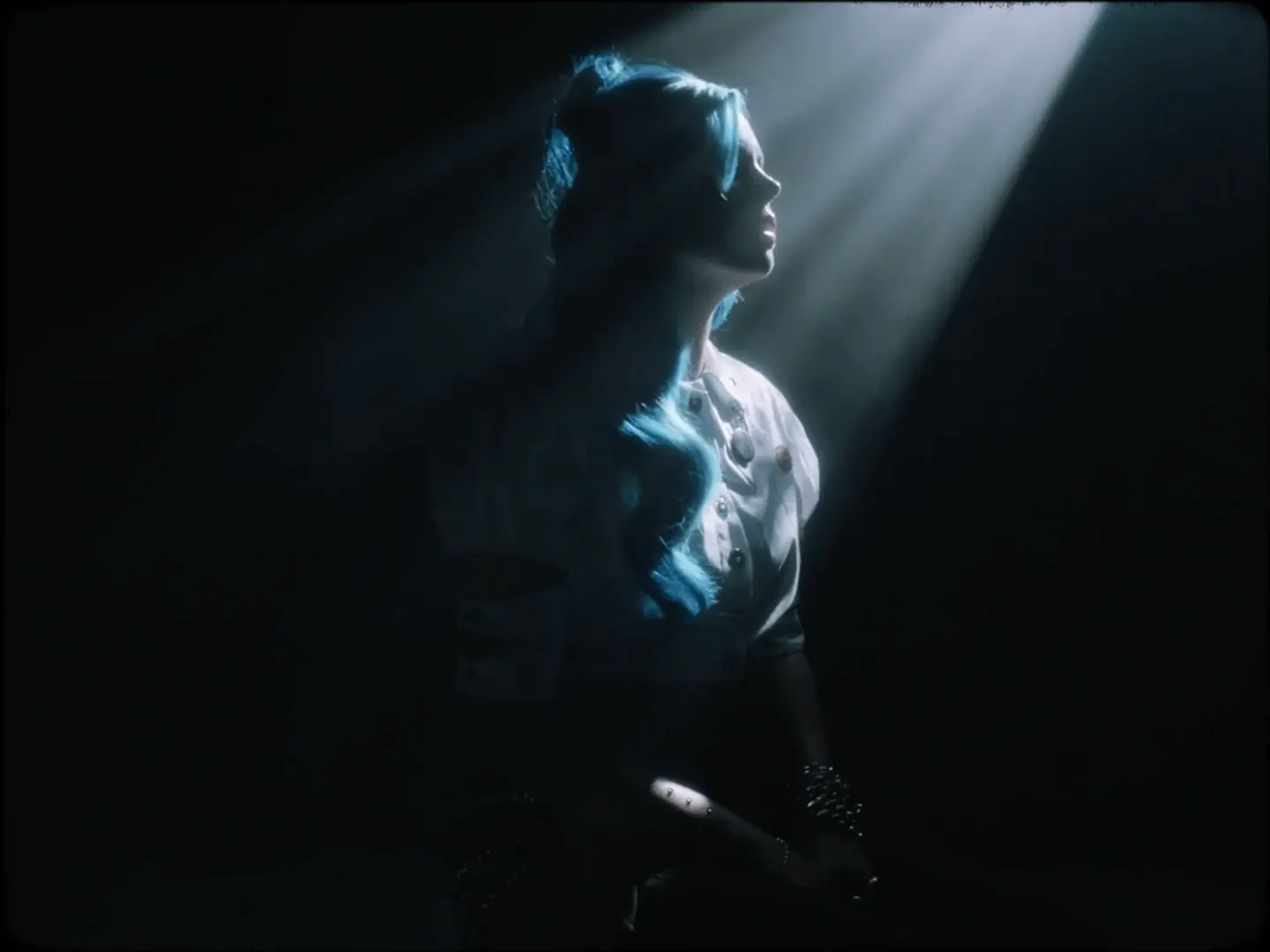 From the 'Ashes By The Morning' music video. Image features artist, Kings Elliot, sitting in a dark room as dappled light from above streams unto her face.