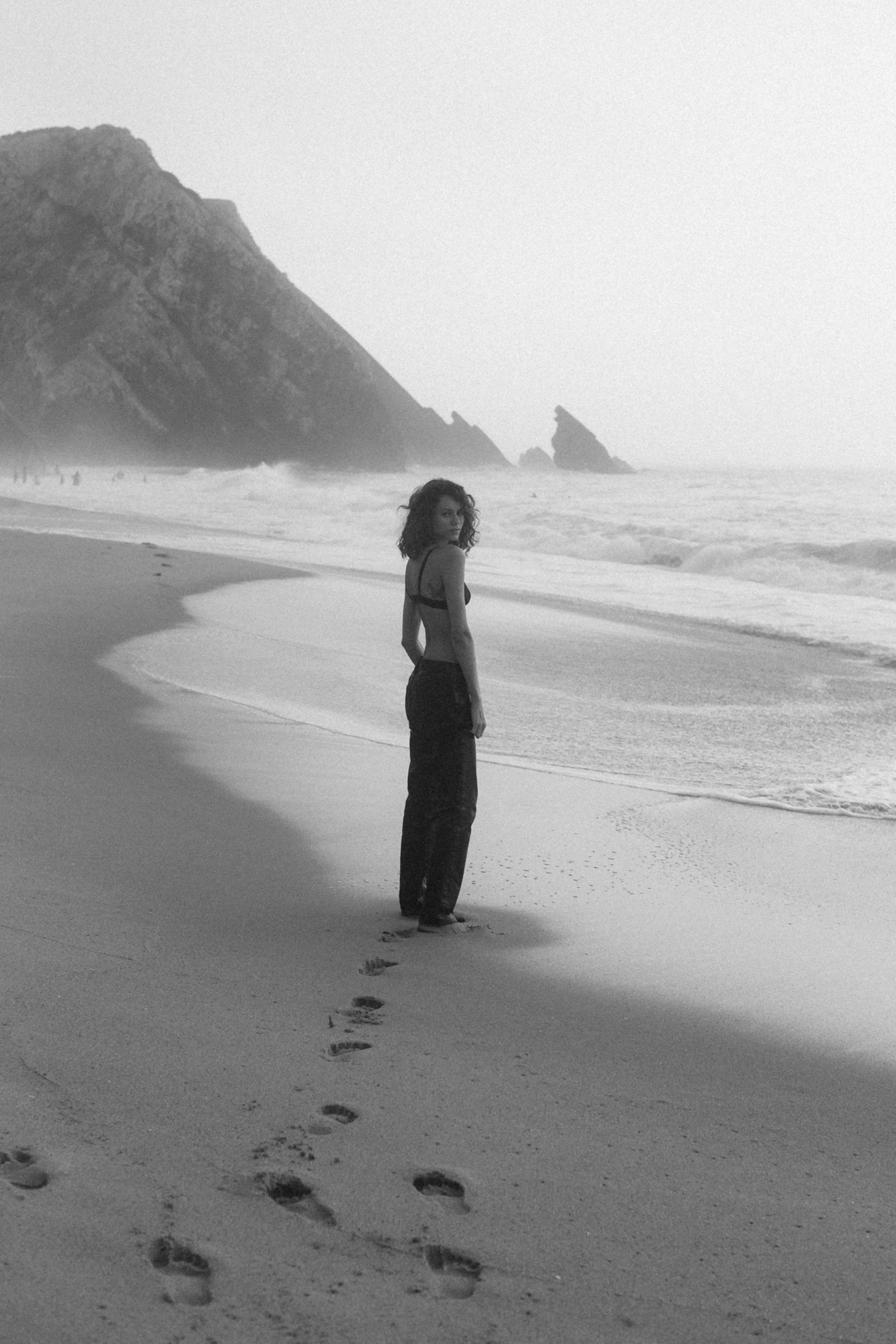 Black and white photograph by Agnes Wonke-Toth. Model, Ketlyn, stands gracefully on the beach near the shoreline, gazing back at the camera.