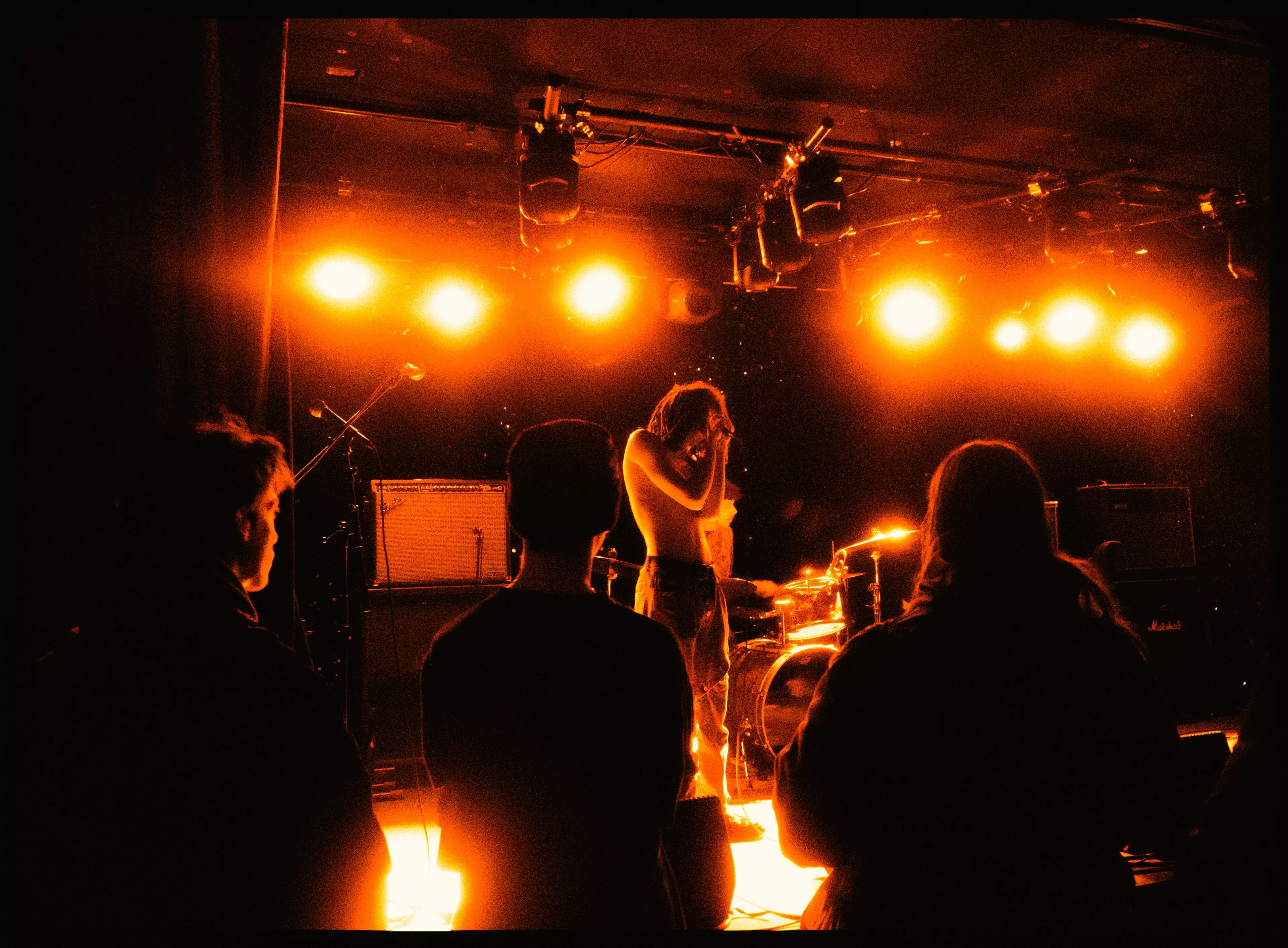 Photograph by Agostina Cerullo. A performer and his band are captured on stage during a gig, illuminated by atmospheric orange stage lights.