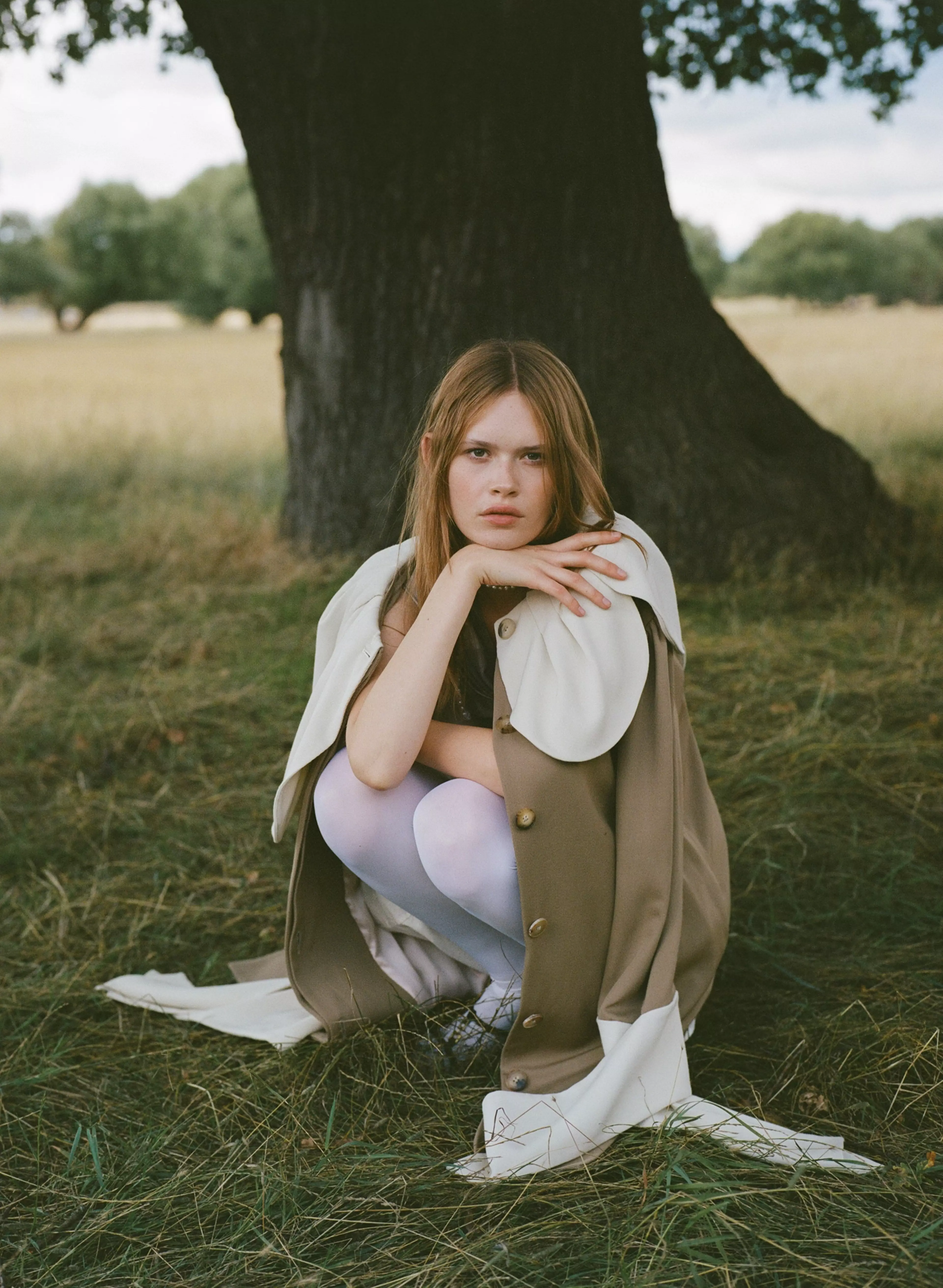 Photograph by Agnes Wonke-Toth. Model Mia crouches in a lush green field, wearing a stylish long coat and facing the camera.