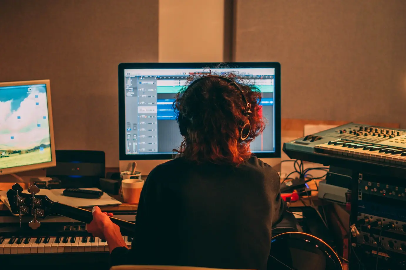 Musician recording a track in a home studio, holding a guitar in front of a keyboard. Image by Maxwell Hunt on Unsplash.